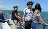 Passengers are enjoying a sunny day on the deck of a boat with a view of the ocean and coastline in the background.