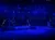 Performers are illuminated with blue and green lighting on a theatrical stage with set elements resembling a nautical theme.