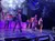 Performers are on stage in dynamic poses amidst theatrical fog and colorful lighting, creating an energetic scene at a live entertainment show.