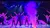 Performers on stage are striking a pose under vivid purple lighting and a fog effect, with a dramatic, futuristic backdrop.