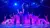 Performers on stage strike a pose under purple and blue lighting, with a dramatic metal structure above them.