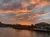 The image captures a breathtaking sunset with vibrant orange and pink hues in the sky, reflecting over a tranquil river beside a neighborhood with elegant waterfront homes.