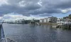 The image captures a cloudy sky over a waterway, with waterfront houses in the distance and the frothy wake of a boat in the foreground.