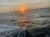 The image captures a serene sunset over a choppy ocean with the sun's reflection creating a golden path on the water, and hints of civilization in the distance.