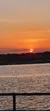 The image captures a serene sunset with a fiery orange sun dipping towards the horizon over a calm body of water, complemented by the silhouette of the distant landscape and a metal railing in the foreground.