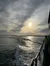 The image captures the view from the side of a boat as it moves across the water, leaving a trail of waves behind, under a partly cloudy sky with the sun hanging low near the horizon.