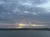 The image depicts a tranquil waterfront scene under a cloudy sky with the sun peeking through the clouds near the horizon.