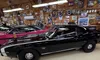 The image shows a black vintage Chevrolet Camaro SS parked in a garage filled with automotive memorabilia and collectibles on the walls.