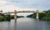 The image shows a bridge spanning over a wide river under a cloudy sky, surrounded by lush greenery.