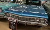 The image shows a classic blue 1966 Chevy Impala on display in a car museum, surrounded by vintage signs and hubcaps.