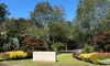 The image shows a landscaped entrance sign for Brookgreen Gardens surrounded by vibrant flowers and trees, under a clear blue sky.
