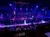 The image shows a live performance in a theater with colorful lighting, a water stage, and a seated audience.