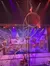 The image shows a performer doing a handstand on a large rotating circular apparatus during a circus act, with a snowy village backdrop and colorful stage lighting.