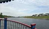 The image shows a river or canal view from a boat with colorful railing, surrounded by houses and a slightly cloudy sky.