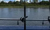 The image shows a serene river landscape viewed through a pair of fishing rods mounted on a boat, under a partly cloudy sky.