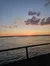 The image shows a serene sunset over a body of water, with clouds in the sky and a railing in the foreground.