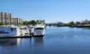 The image shows a serene waterfront scene with boats docked near a residential area and a bridge in the background, under a clear blue sky.