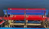 The image shows a vibrant red and blue paddle wheel of a riverboat against a backdrop of dark, rippling water.