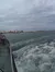The image shows a view from a boat with swirling water in the foreground and a distant shoreline with buildings and a pier under a cloudy sky.