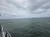 The image shows an overcast sky above a calm sea, taken from the side of a boat with part of the railing visible and a person seen in the corner looking out over the water.