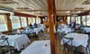 The image shows the interior of a wooden restaurant or café on a boat, with several tables covered in white tablecloths and a few people seated along the side.