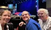 Three people are smiling and posing for a photo inside a theater or auditorium, with a stage and lighting setup visible in the background.