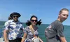 Three people are standing on a boat with the ocean in the background, with two of them wearing sunglasses and one donning a hat, as the wind seems to be blowing their hair.