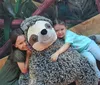Two children are happily hugging a large stuffed sloth while sitting on a wooden bench with a leafy background.