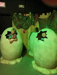 Two children are playfully posing inside large, cracked dinosaur egg replicas in a jungle-themed setting.