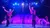 Two performers in matching red and black costumes are taking a bow on a brightly lit stage with other performers in the background.