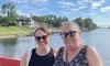 Two smiling women are enjoying a sunny day aboard a boat with a scenic view of the water and houses in the background.