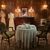 The image shows an ornately decorated room with a vintage dining setup, two mannequin-dressed in period clothing, and a portrait hanging above a wooden sideboard, creating an ambiance reminiscent of a past era.