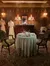 The image shows an ornately decorated room with a vintage dining setup, two mannequin-dressed in period clothing, and a portrait hanging above a wooden sideboard, creating an ambiance reminiscent of a past era.