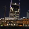 The image features a night view of a brightly lit, distinctive high-rise building in an urban setting, juxtaposed with older, historic architecture in the foreground.