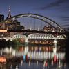 The image shows a striking nighttime cityscape featuring illuminated bridges over a calm river with reflections of city lights on the water.