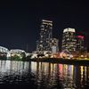 The image captures a vibrant cityscape at night, with illuminated skyscrapers reflecting off the water's surface, and a lit bridge to the left.