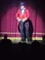 A person in a red and black ringmaster costume stands on stage before an audience, slightly blurred implying movement or a low-light capture.