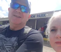 A man and a woman take a close-up selfie with the man wearing sunglasses and a cap, while the background shows a building with the sign 