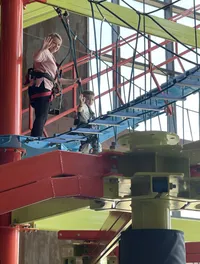 Two individuals, likely a woman and a child, are walking across a colorful indoor ropes course while wearing safety harnesses and are smiling and waving at the camera.