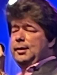 A man in a suit is passionately singing into a microphone on stage with blue stage lights in the background.