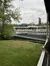 A large multi-deck riverboat with paddle wheels is moored beside a green, tree-lined riverbank, viewed from a balcony with metal railing.
