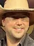 A person wearing a cowboy hat and holding a guitar is posing in front of a backdrop that reads 