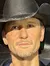 A wax figure or life-sized statue of a man wearing a cowboy hat, casual jacket, and jeans poses with one arm extended, in what appears to be a staged backstage area with images of people cheering in the background.