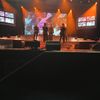 A group of performers is standing on stage with coordinated outfits, entertaining an audience, with colorful stage lighting and background screens displaying content related to the performance.