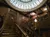 The image shows an ornate wooden staircase with intricate railings, a decorative statue holding a lamp, and a large geometric skylight above.