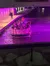 A group of people enjoy a boat tour under a vividly lit purple underpass on a riverwalk at night.