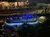 A boat adorned with blue lights carries passengers along a river at night, with spectators on the riverside walkway.