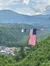 The image shows a large American flag hanging from a suspension bridge with multiple flags, overlooking a lush green landscape and a small town nestled in the valley below.