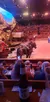 The image shows a live horse-drawn wagon performance in an indoor arena with an audience watching from tiered seating and a bottle of beer in the foreground.