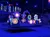 The image shows a vibrant stage adorned with illuminated, colorful artificial flowers, set against an audience in a dimly lit arena-like setting.